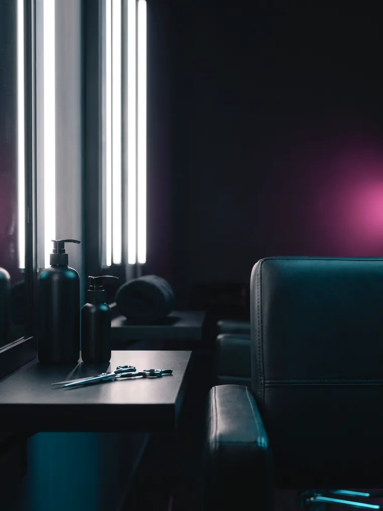 One styling station close to: pump bottles and open shears on a dark counter, a rolled towel behind them, a leather chair at the edge of the frame.