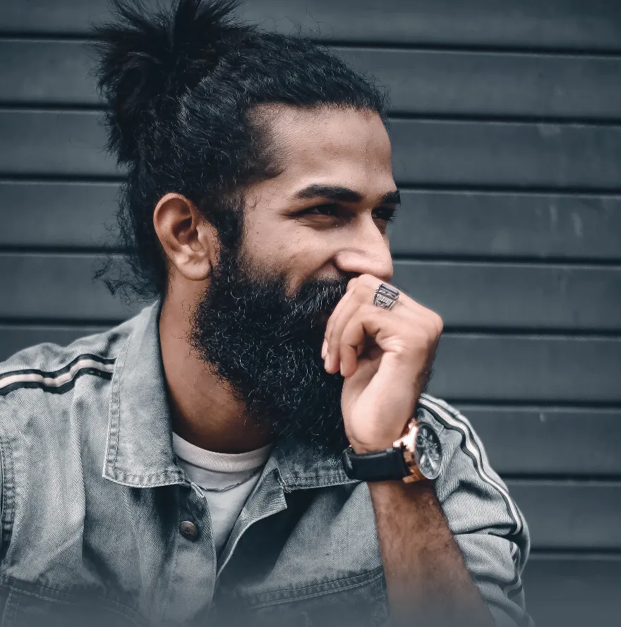 A bearded man laughing with one hand to his face, in a denim jacket against a dark grey shutter.